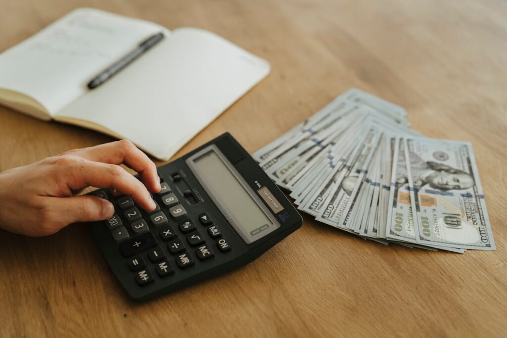 Hand on calculator next to a stack of hundred-dollar bills and a notebook