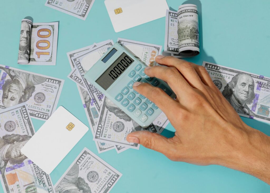 Hand typing costs into a calculator surrounded by hundred-dollar bills